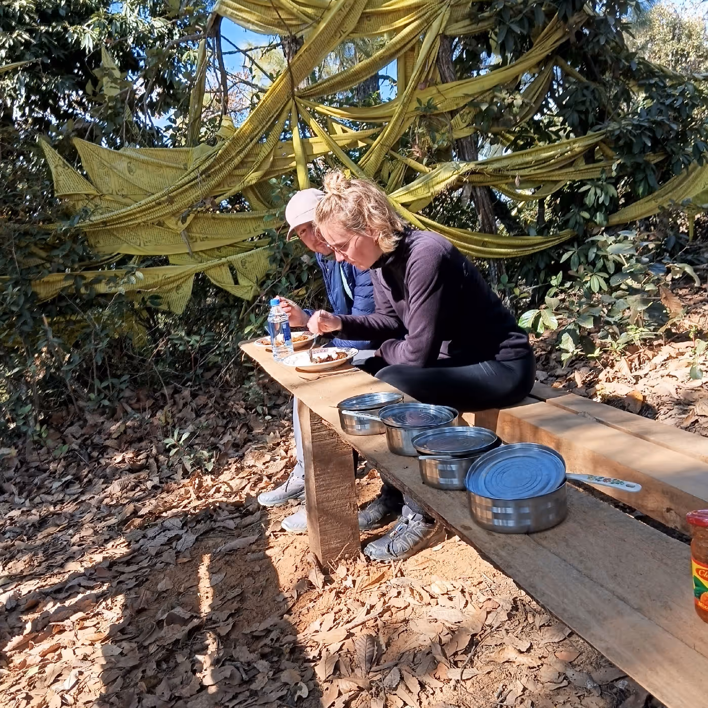 Two trekkers enjoy a rustic lunch on a wooden bench under fluttering prayer flags, surrounded by forest and dry leaves along the Bumdra trail.