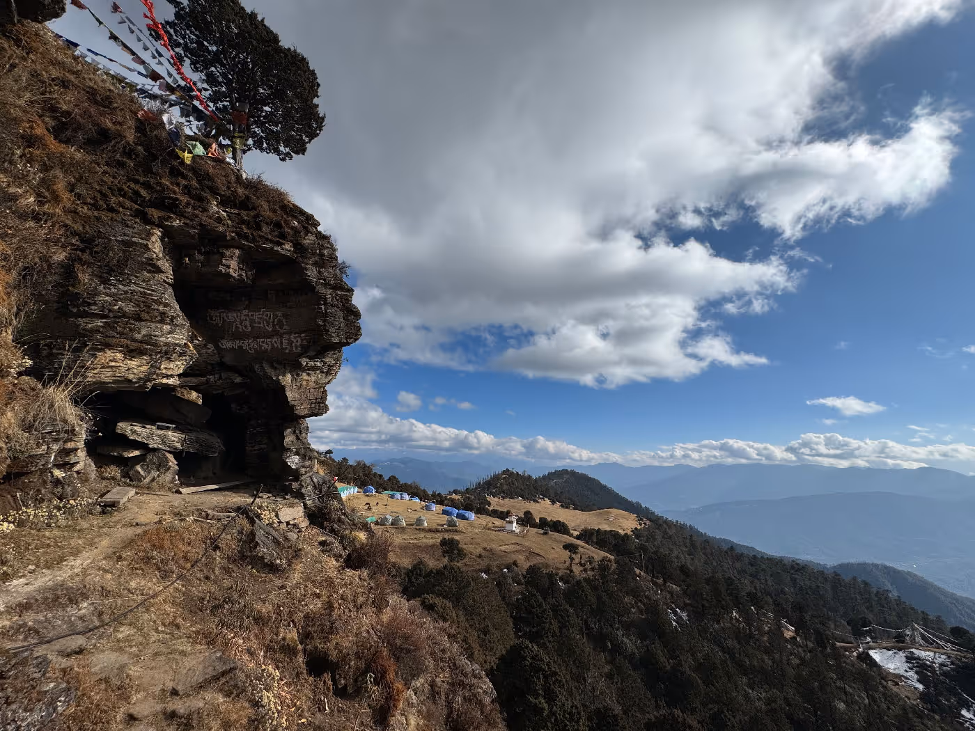 View from a rocky cliffside cave etched with Buddhist inscriptions, overlooking a serene meadow dotted with colorful camping tents and prayer flags, with pine forests and layers of mountains stretching beyond.