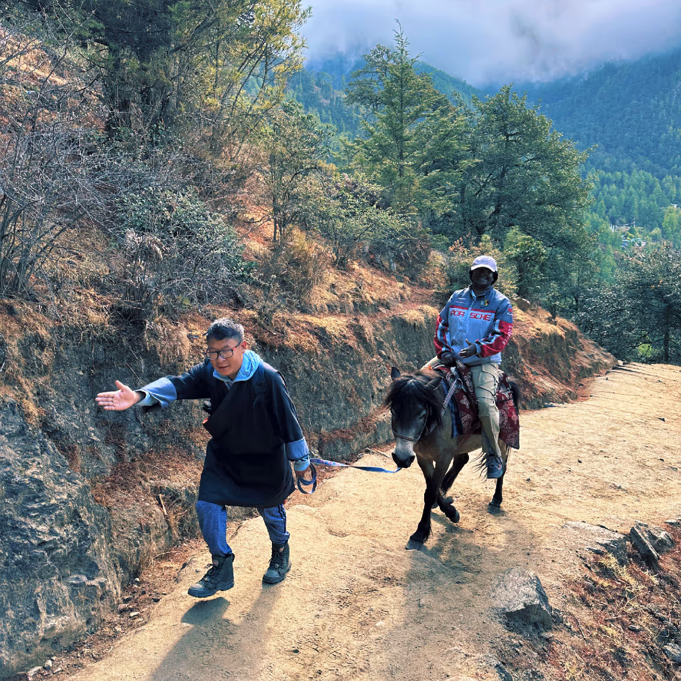 A cheerful traveler rides a pony led by a local guide along the dusty forest trail to Tiger’s Nest, with early morning mist veiling the distant hills.