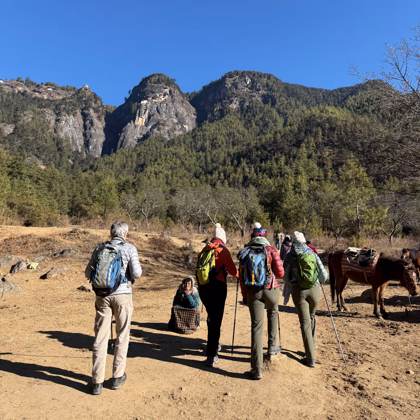 A group of travelers stand at the base of a forested mountain near Tiger’s Nest, chatting with a local woman seated on the trail, while horses wait nearby.