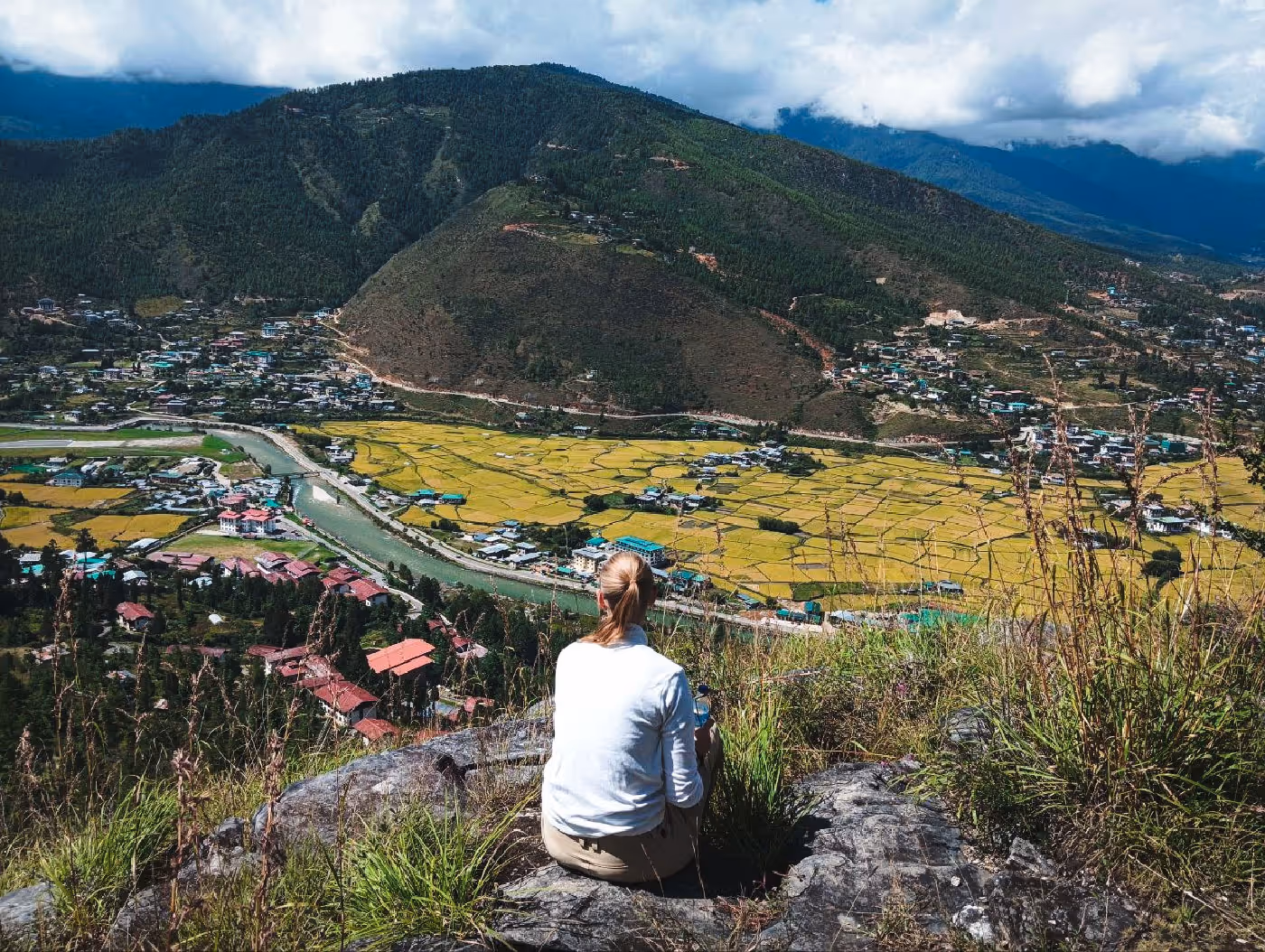 Solo hiker gazes over Paro’s golden rice fields and glistening river from a serene viewpoint along the Zuri Dzong hike in Bhutan.