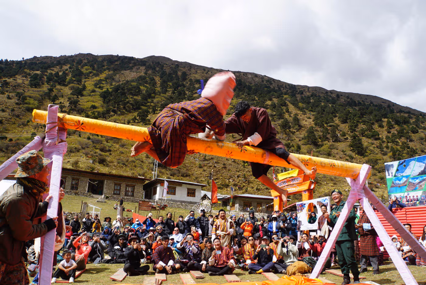 Locals playfully competing on a padded pole during the Jumolhari Mountain Festival, with a cheering crowd and highland backdrop.