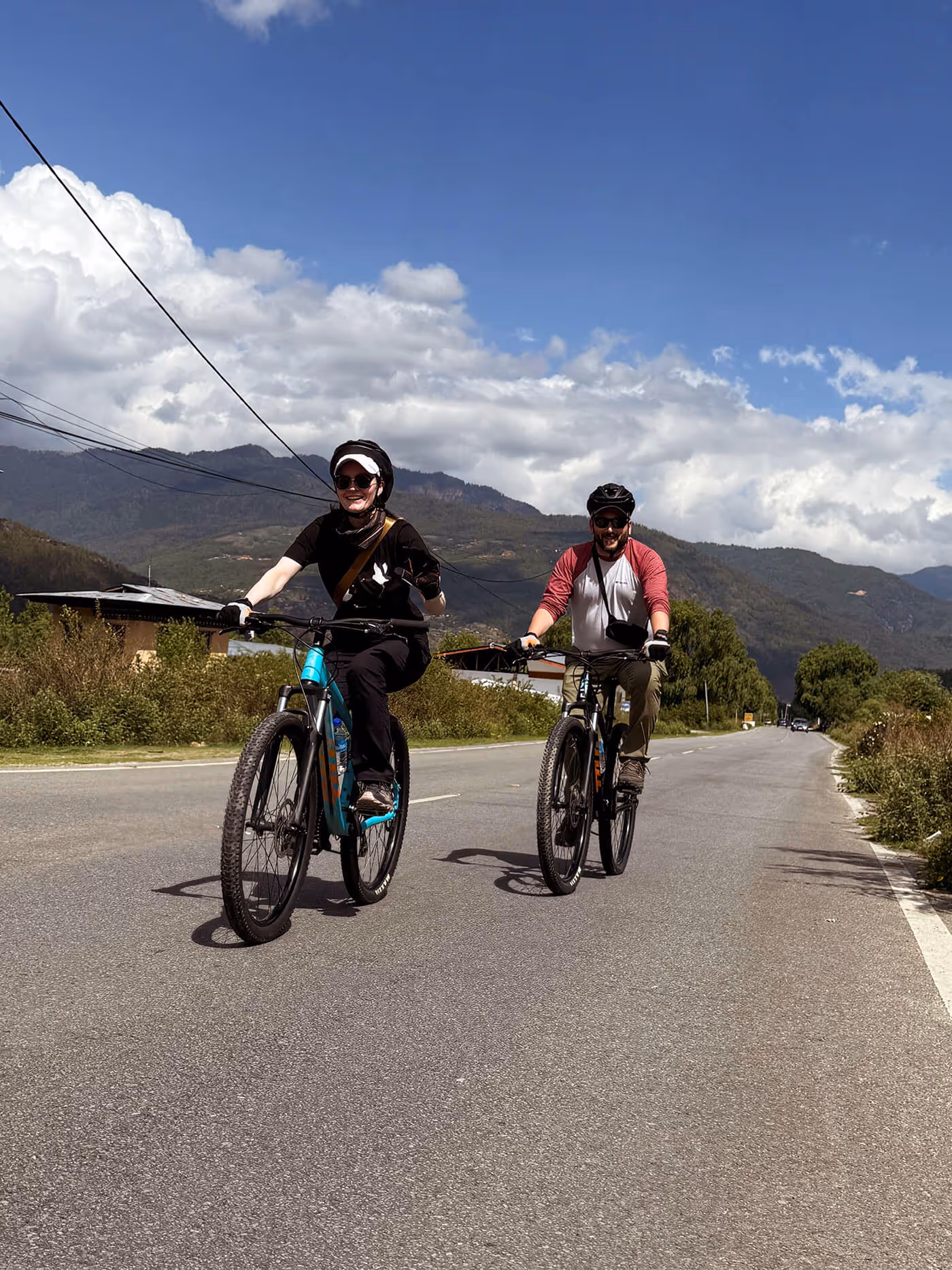 A couple cycles side by side on a quiet mountain road, surrounded by open skies and forested peaks, capturing the carefree spirit of a Bhutan adventure.