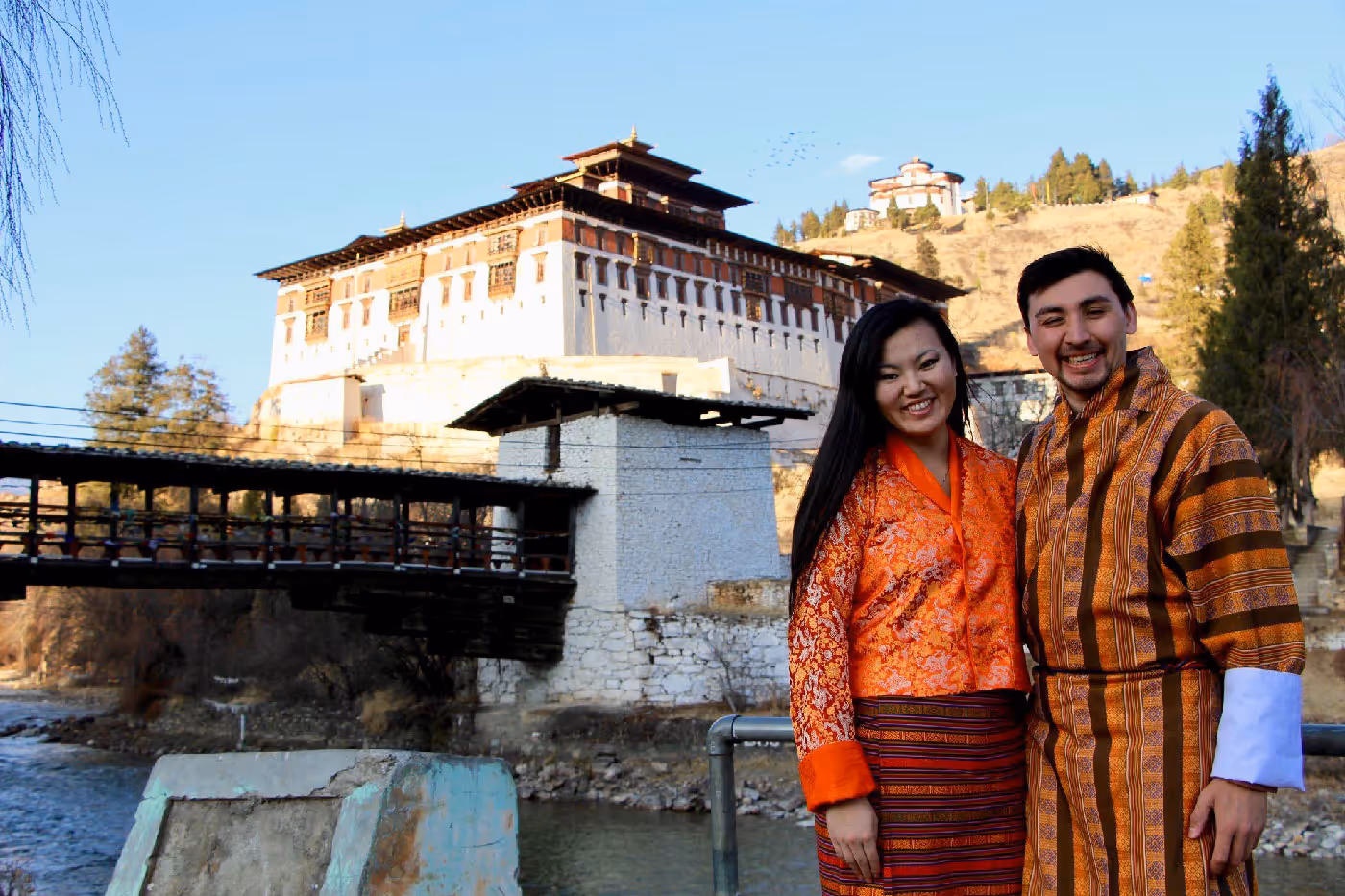 A smiling couple wearing kira and gho stands by the river with the majestic Paro Rinpung Dzong rising behind them at golden hour.