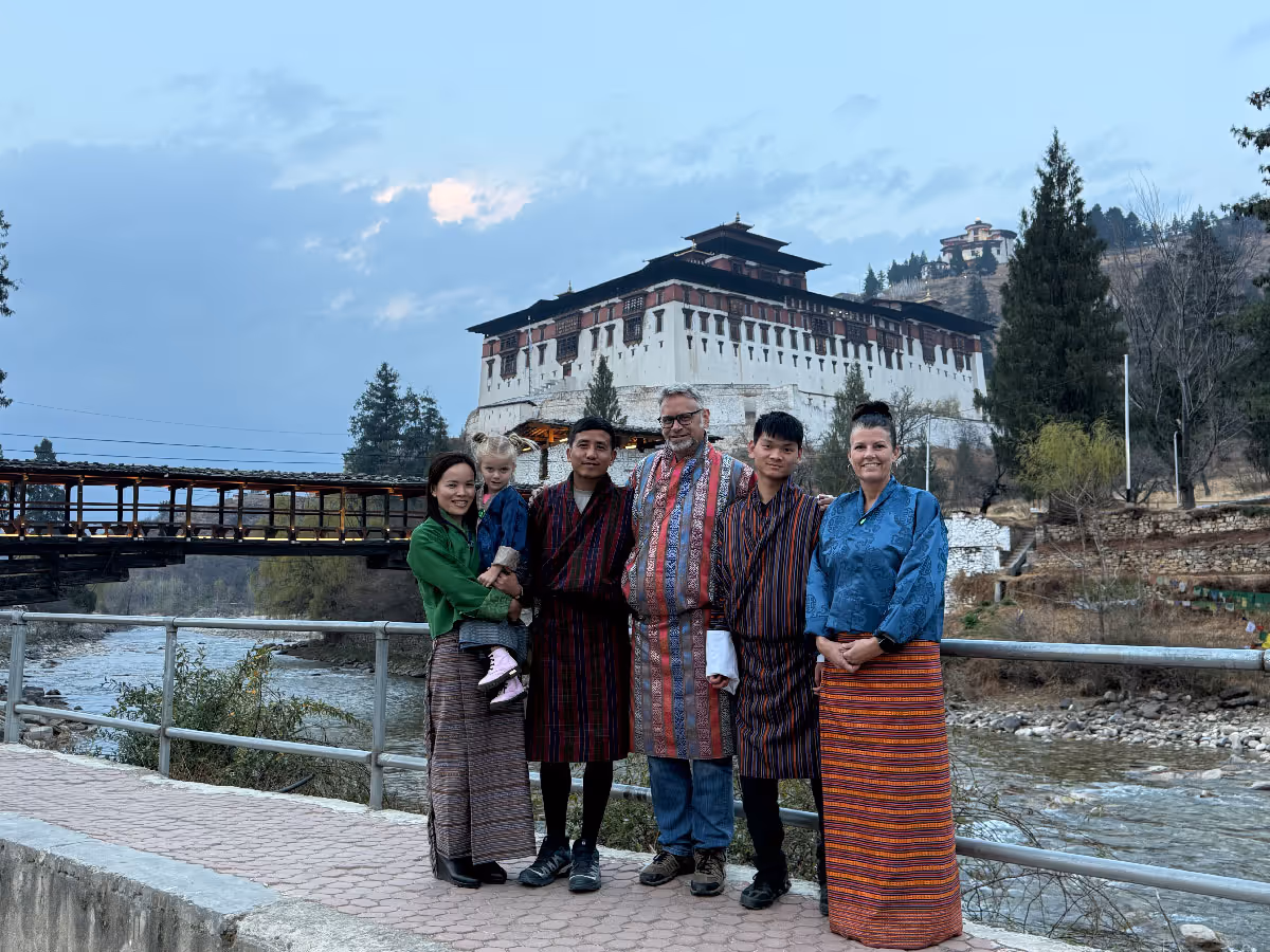 A family group stands warmly together along the riverside, dressed in gho and kira, with Paro Dzong and the traditional cantilever bridge in the backdrop.