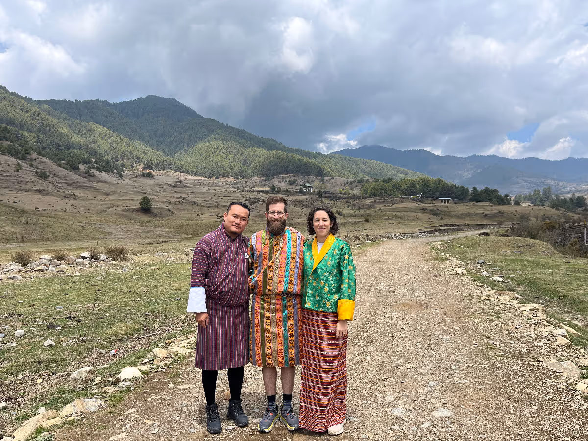 Two international travelers and a local guide pose against a dramatic highland landscape in Bhutan, dressed in traditional Bhutanese clothing.