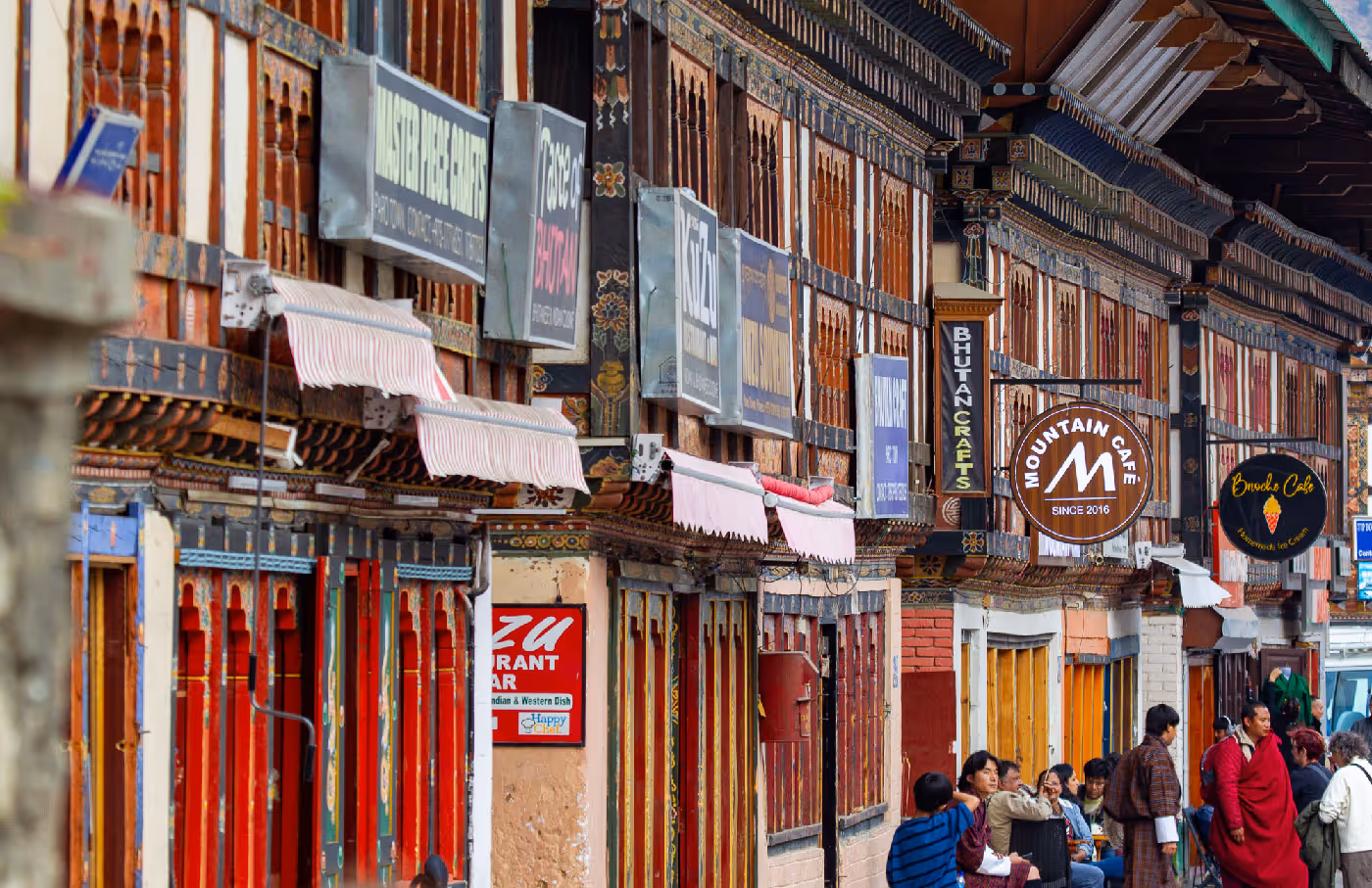 Intricately painted Bhutanese buildings lined with restaurants, cafés, and craft shops in the heart of Paro town, with locals passing by under striped awnings.