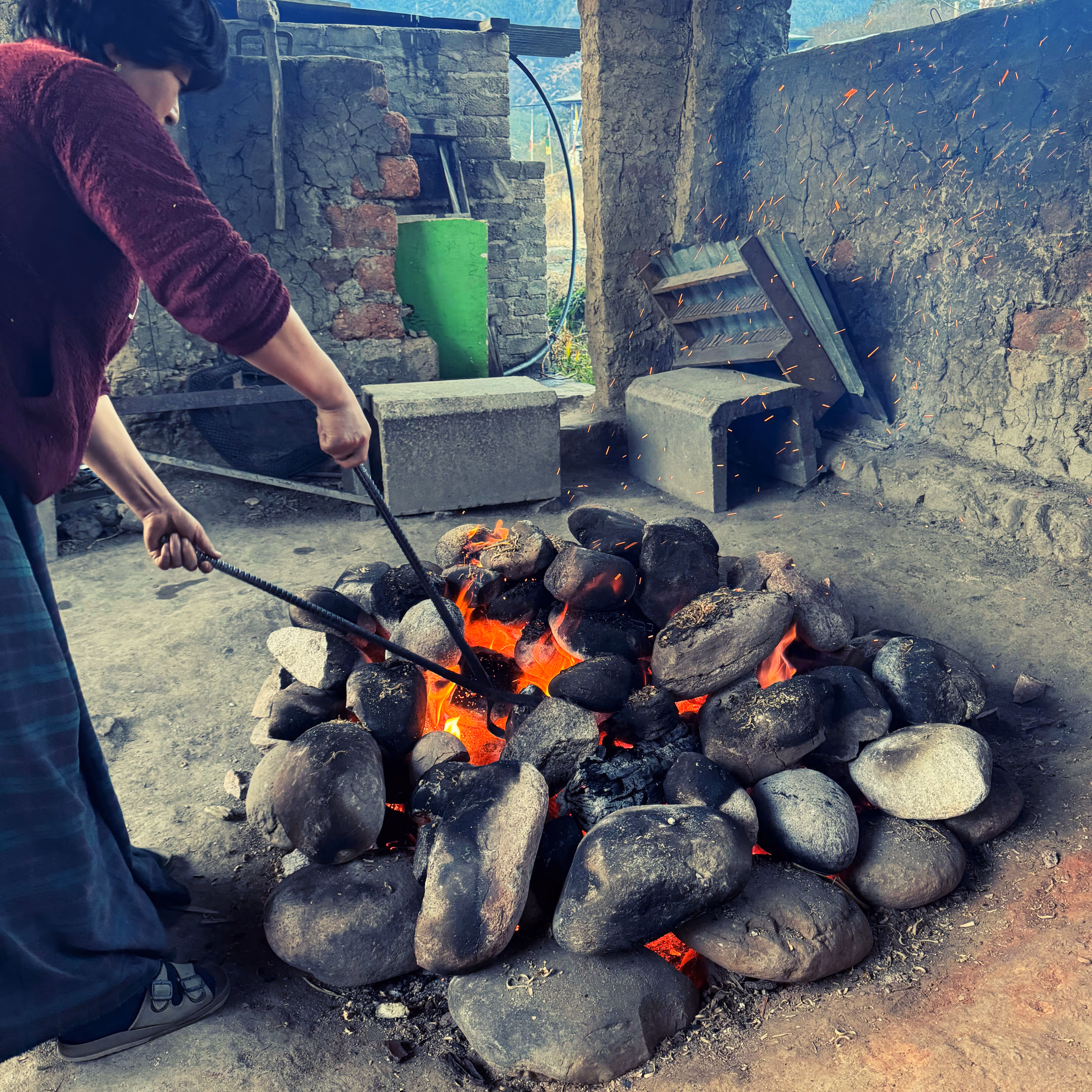 Bhutanese woman using iron tongs to heat river stones for a traditional hot stone bath.