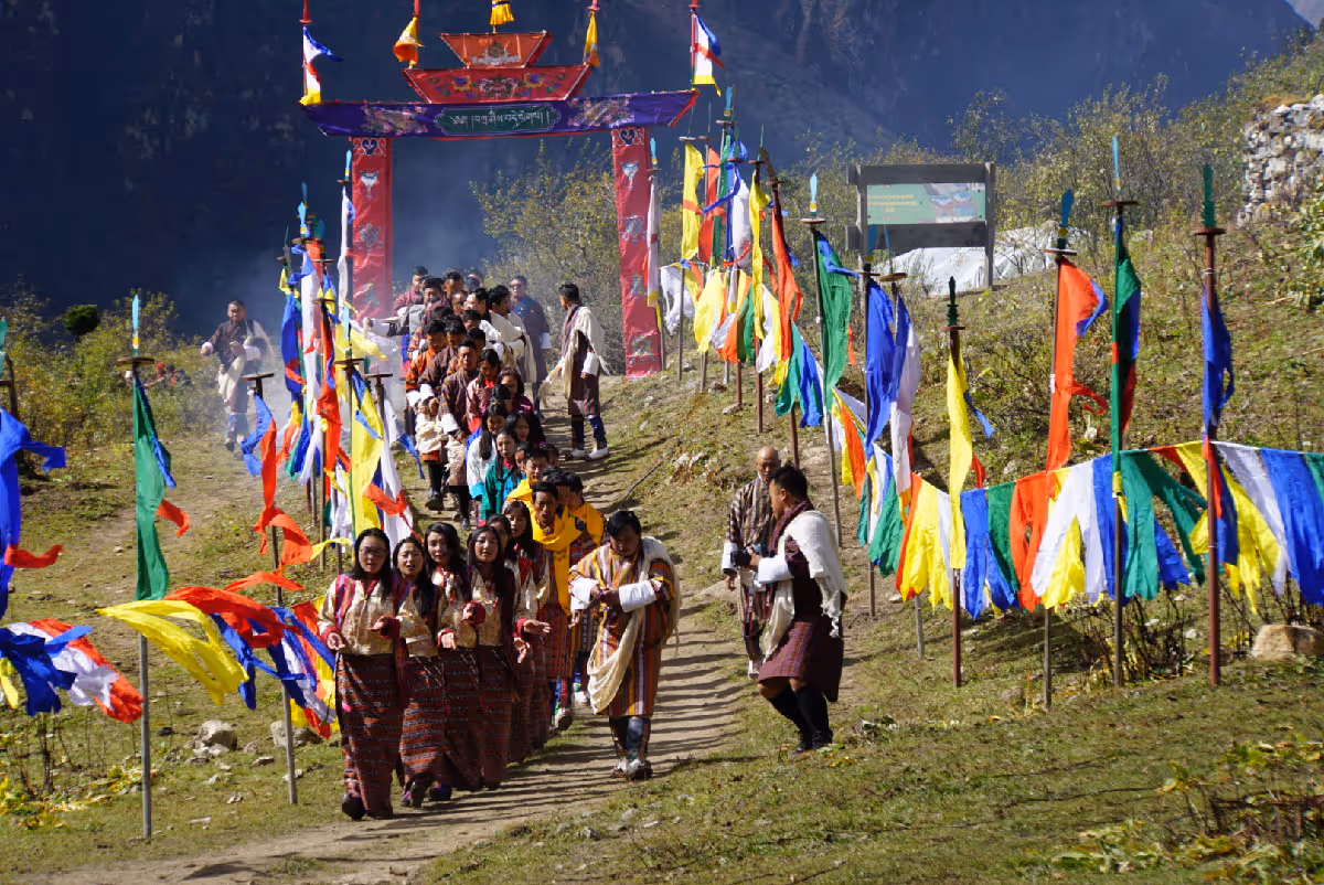 A ceremonial procession of performers and villagers dressed in traditional attire walks through fluttering prayer flags under a festive arch at Jumolhari Mountain Festival.