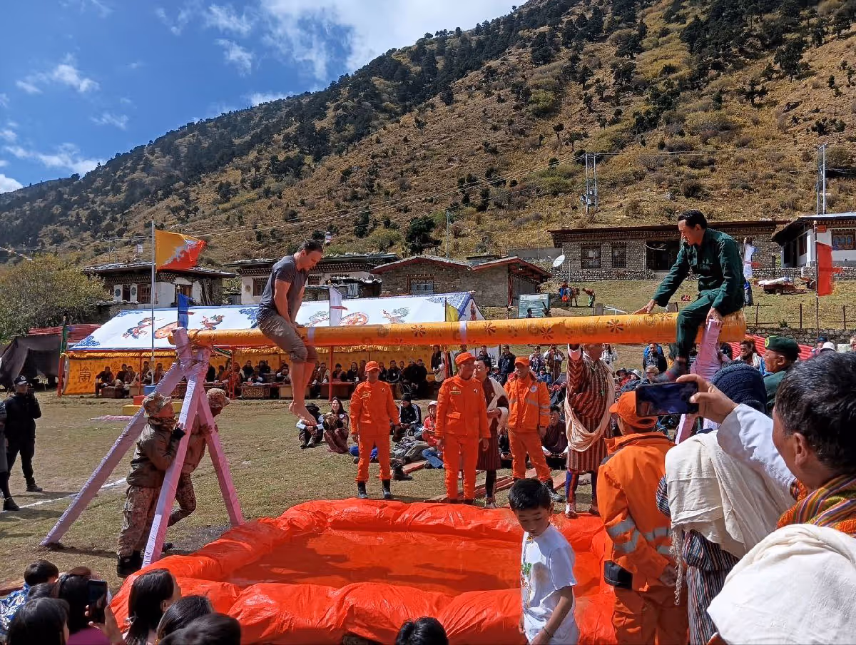 A guest and local participant balance on a traditional pole game above a red landing mat, surrounded by curious onlookers and uniformed marshals at the Jumolhari Mountain Festival.