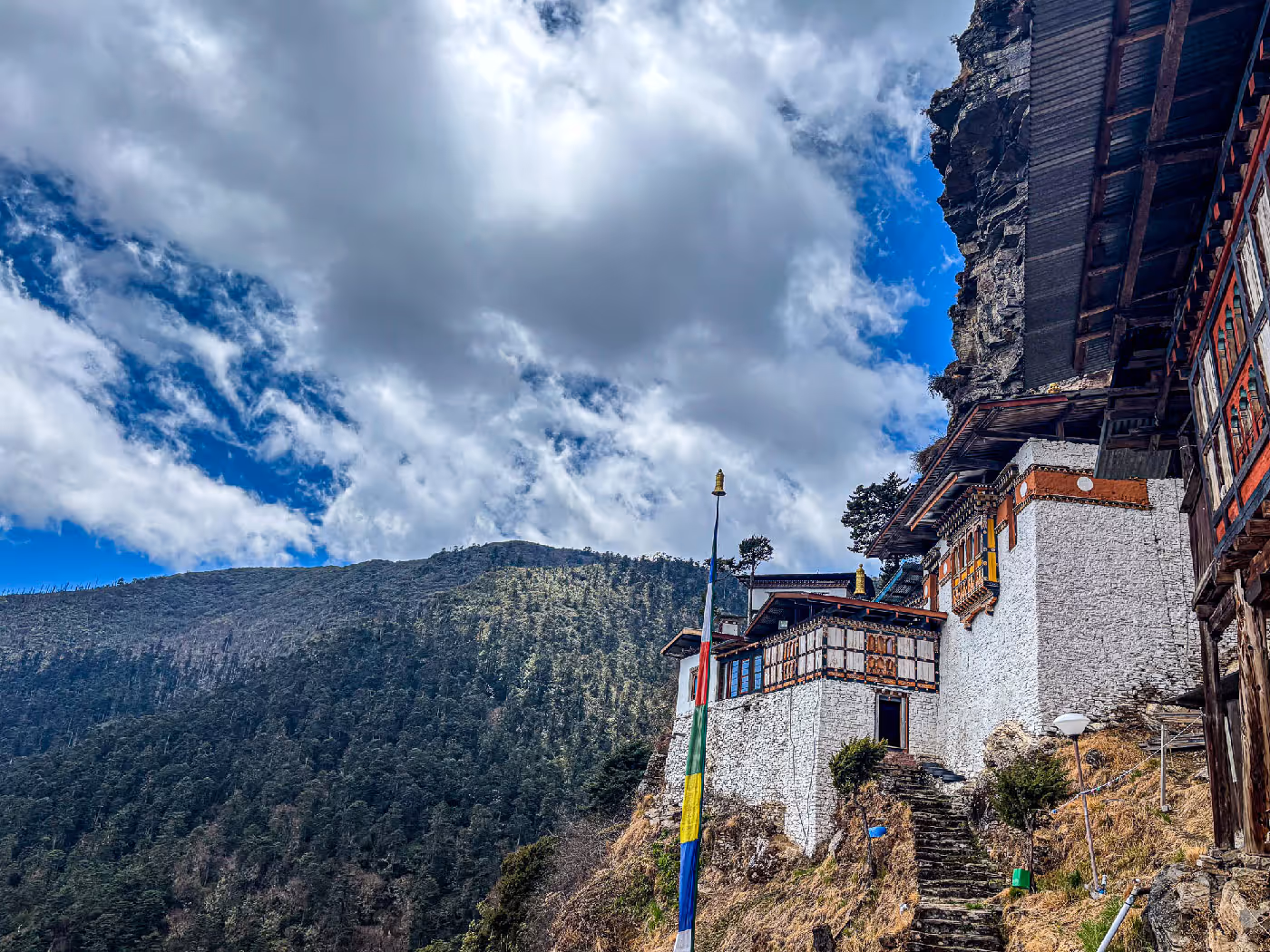 Traditional Bhutanese nunnery perched on a cliffside with fluttering prayer flags and pine-covered mountains in the background under a dramatic sky.