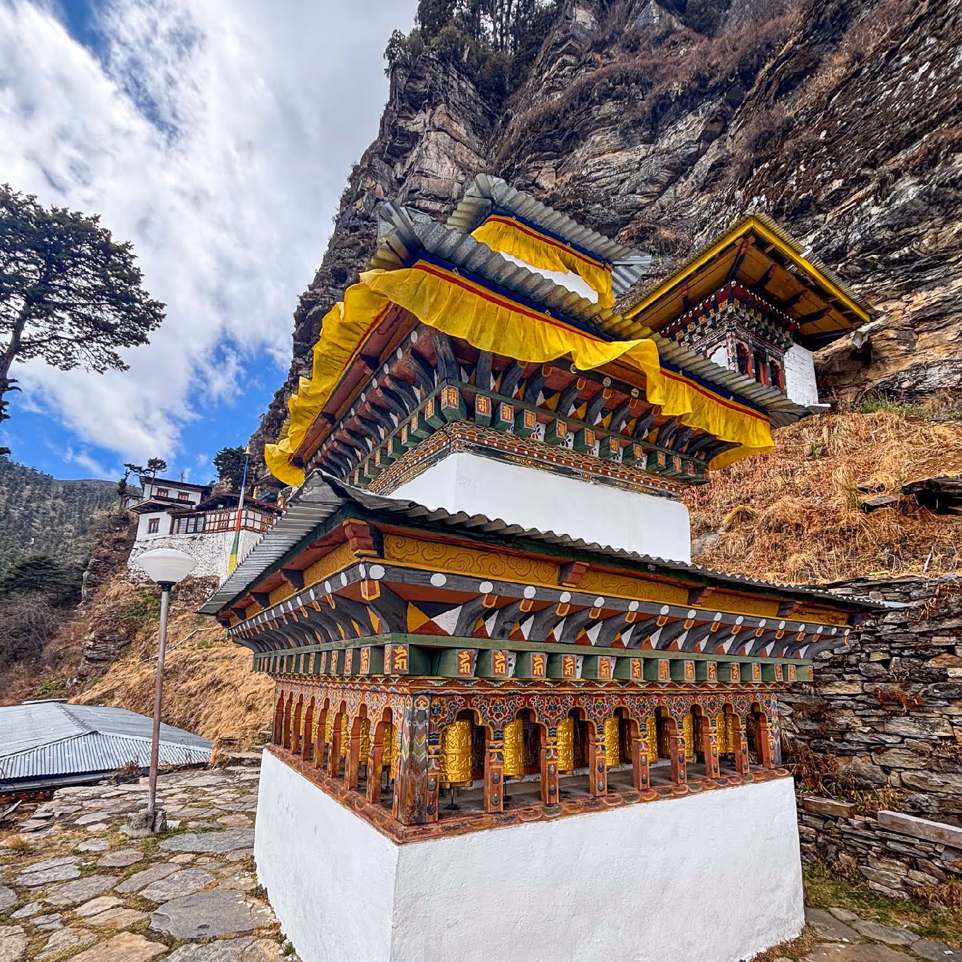 Intricately carved white and gold stupa-style prayer wheel structure nestled against a rocky cliff, with layers of spinning wheels at the base.