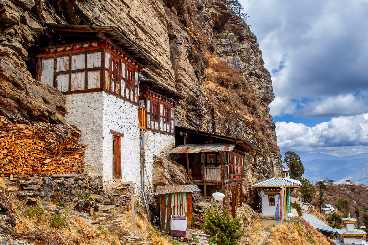 Cluster of rustic meditation huts and traditional monastic dwellings built into the cliff face, surrounded by prayer flags and simple pathways.