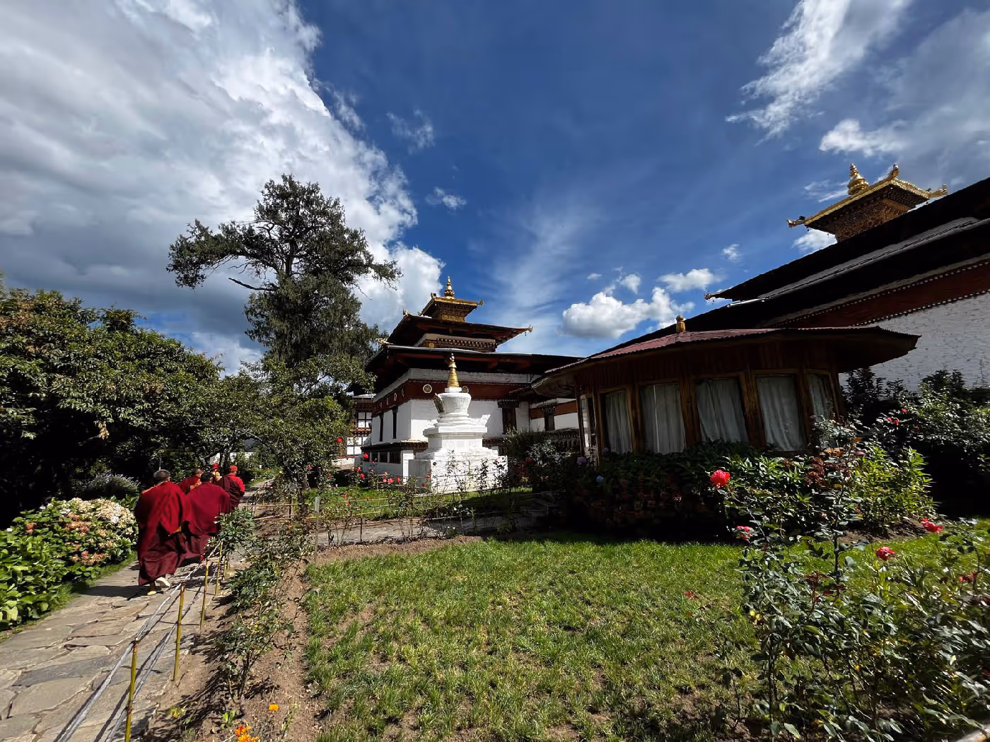Monks walking past Kyichu Lhakhang’s central chorten, with vibrant rose gardens and whitewashed walls glowing under bright summer skies.