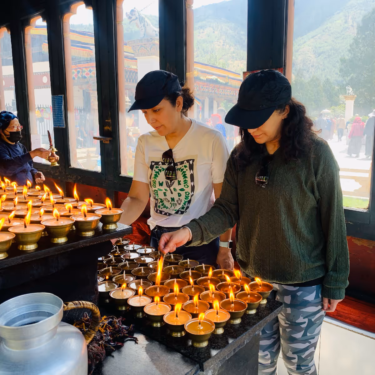 Two guests light rows of golden butter lamps inside a serene Bhutanese temple, bathed in soft morning light and the warmth of flickering flames.