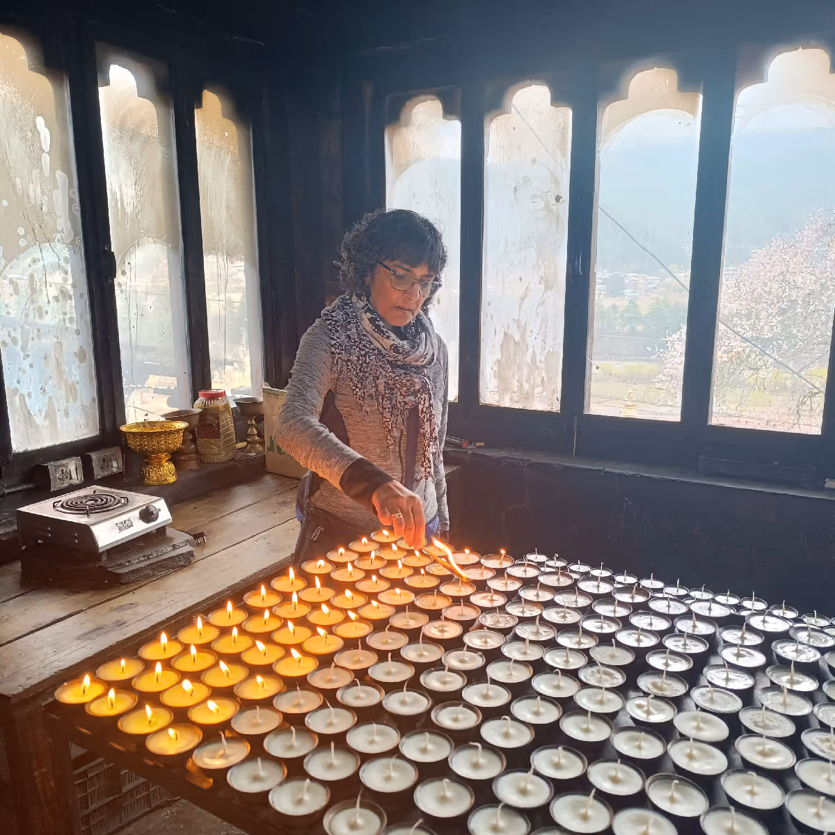 A solo guest lights dozens of butter lamps in a peaceful corner of a monastery, with panoramic valley views framed by arched windows.