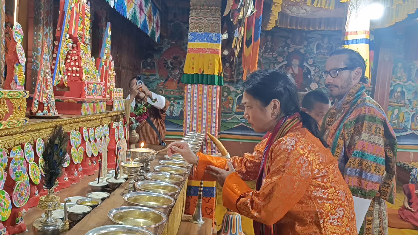 Guest in a bright kira places a water offering inside a vibrant Bhutanese temple, surrounded by intricate murals and sacred relics.