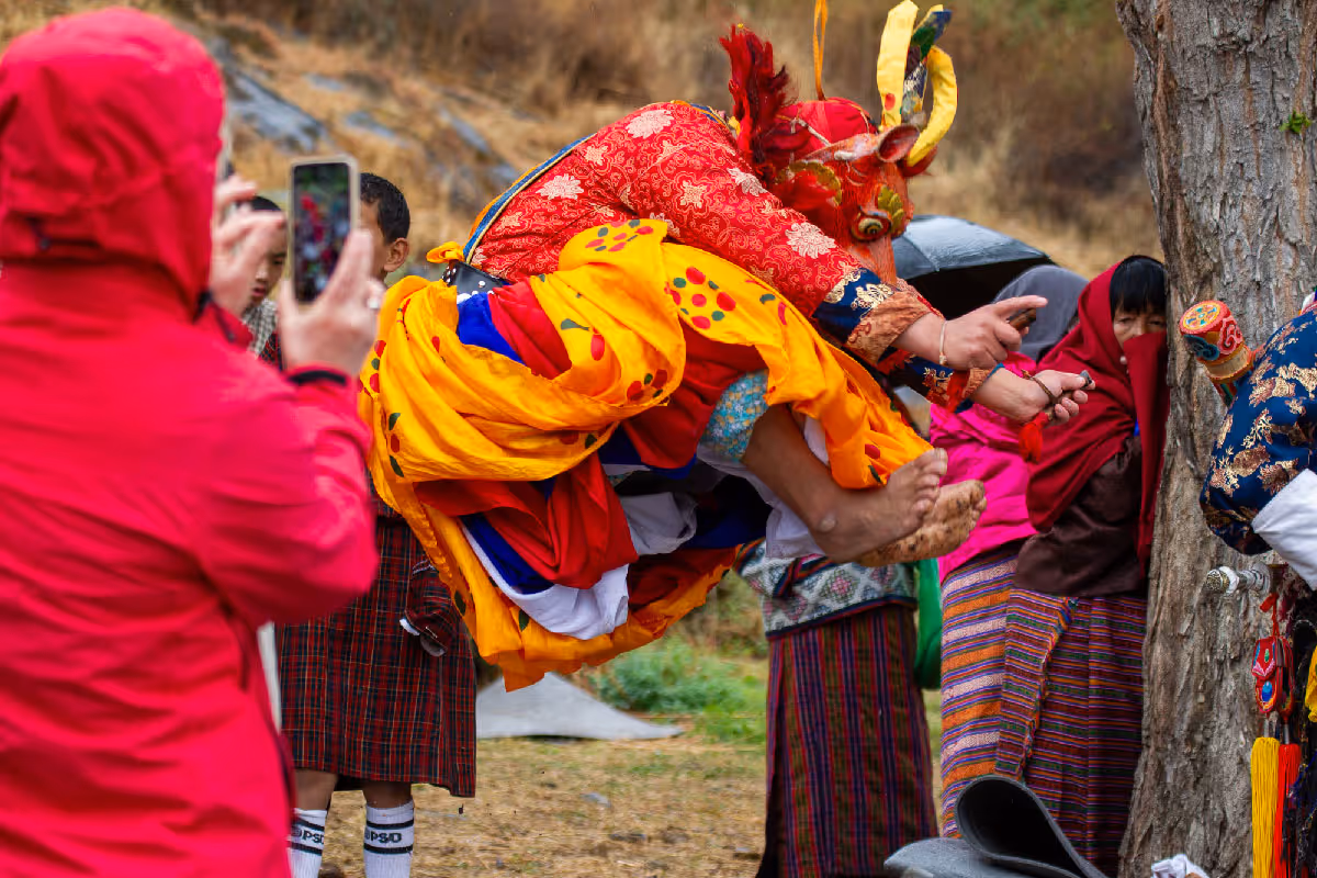 A guest and local participant balance on a traditional pole game above a red landing mat, surrounded by curious onlookers and uniformed marshals at the Jumolhari Mountain Festival.