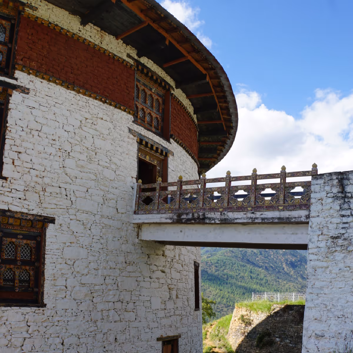 Close-up view of a decorated bridge connecting the upper levels of the Paro National Museum, with intricate woodwork and distant forested hills visible below.