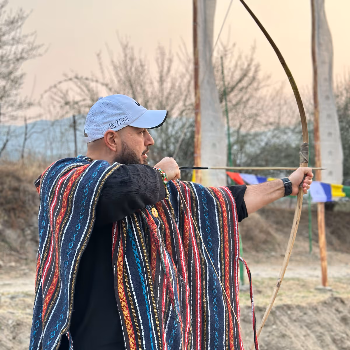 Guest in traditional striped fabric poncho draws a bamboo bow during an archery session in Bhutan.