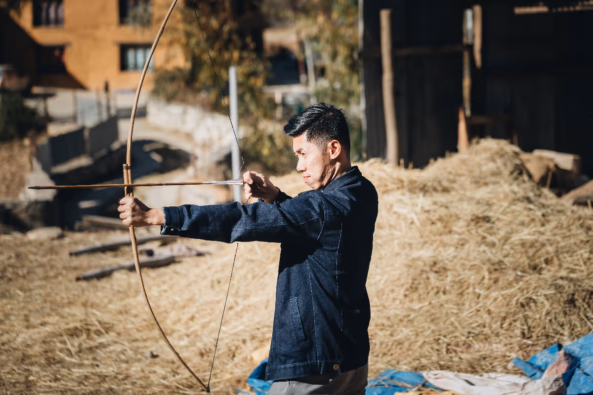 Bhutanese man in a denim jacket focuses intently as he draws a traditional bamboo bow.