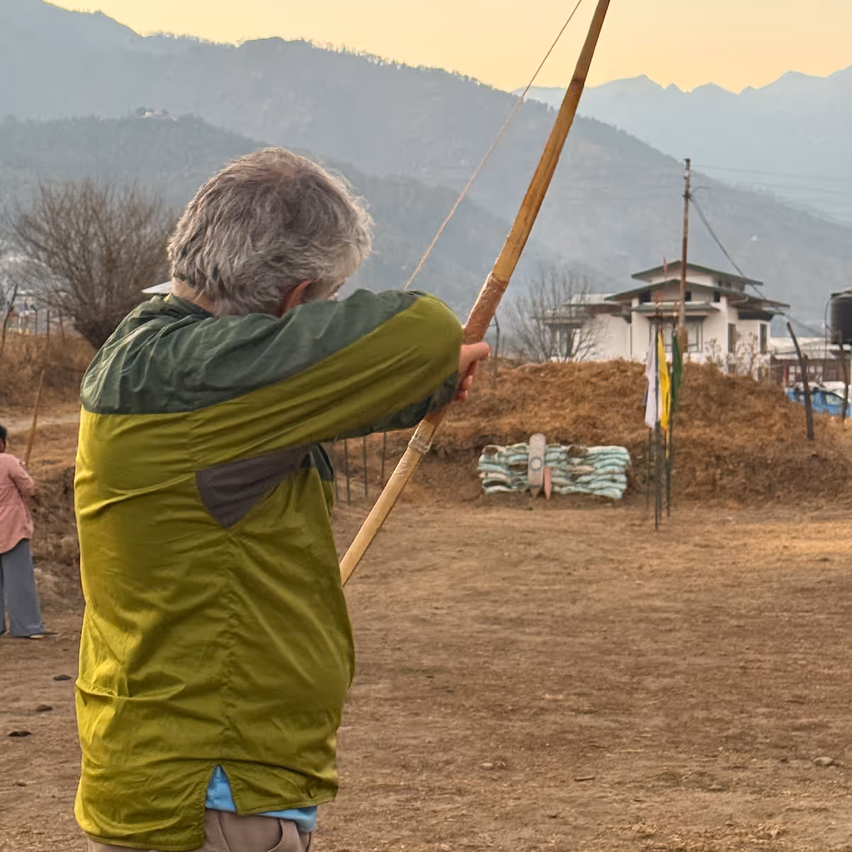 International guest in a green windbreaker practices Bhutanese archery against a scenic mountain backdrop.