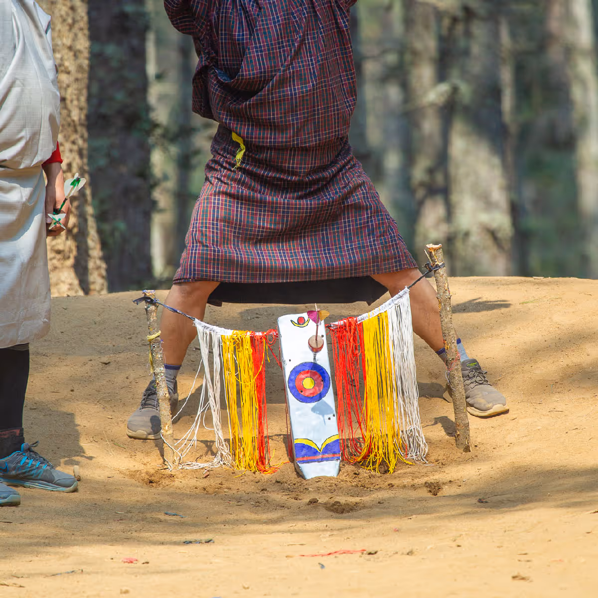 Close-up of a Bhutanese dart target adorned with colorful threads in a sandy forest clearing.