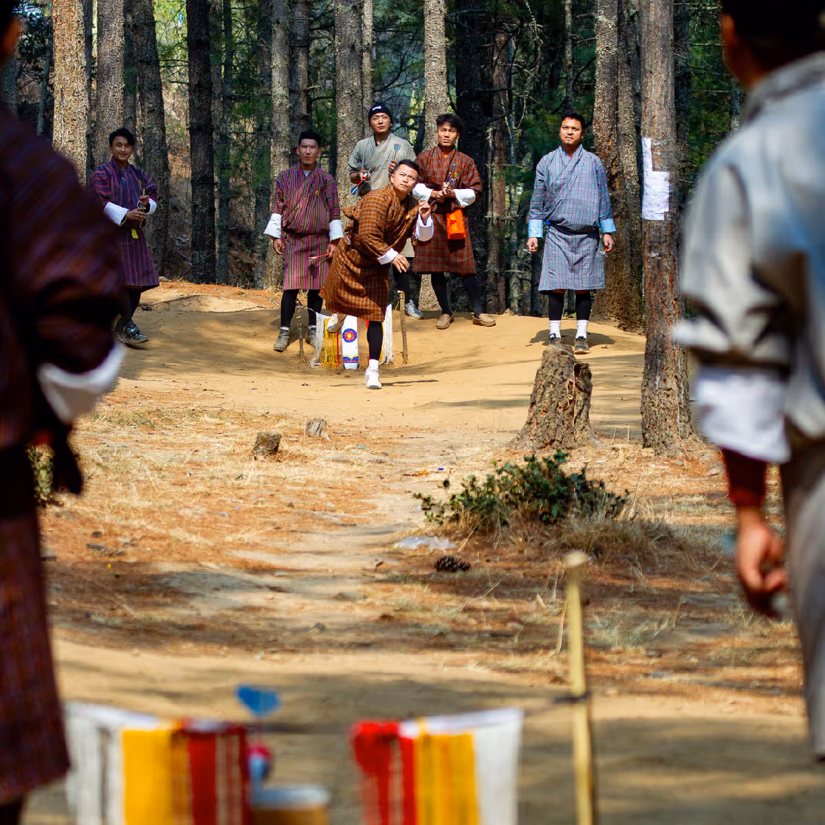 Young Bhutanese men in gho play a traditional dart game in the forest, mid-throw captured in motion.