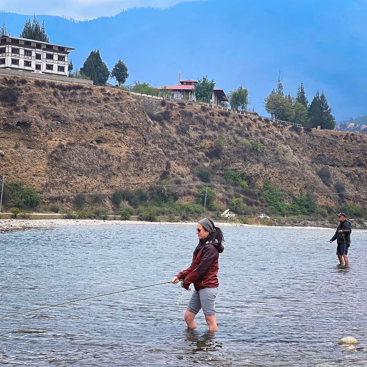 A woman stands mid-river fly-fishing near traditional Bhutanese homes perched atop a dry, rocky slope under a clear sky.