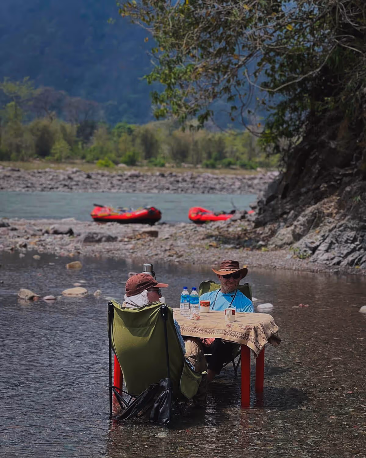 Two travelers relax at a table set in shallow river water, surrounded by nature, with red rafts resting on the riverbank behind them.