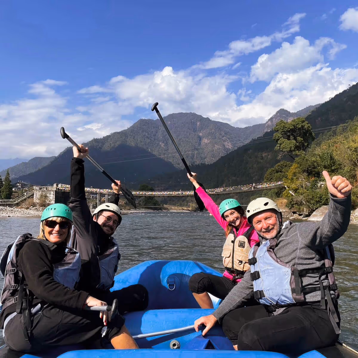 Smiling group rafting on the Punakha River in Bhutan, with a prayer-flag-draped suspension bridge behind.