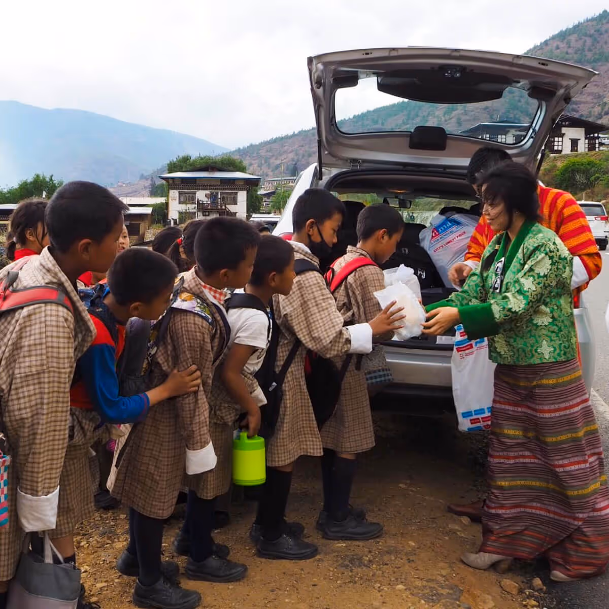 Bhutanese schoolchildren receiving packed gifts from volunteers outside a vehicle.