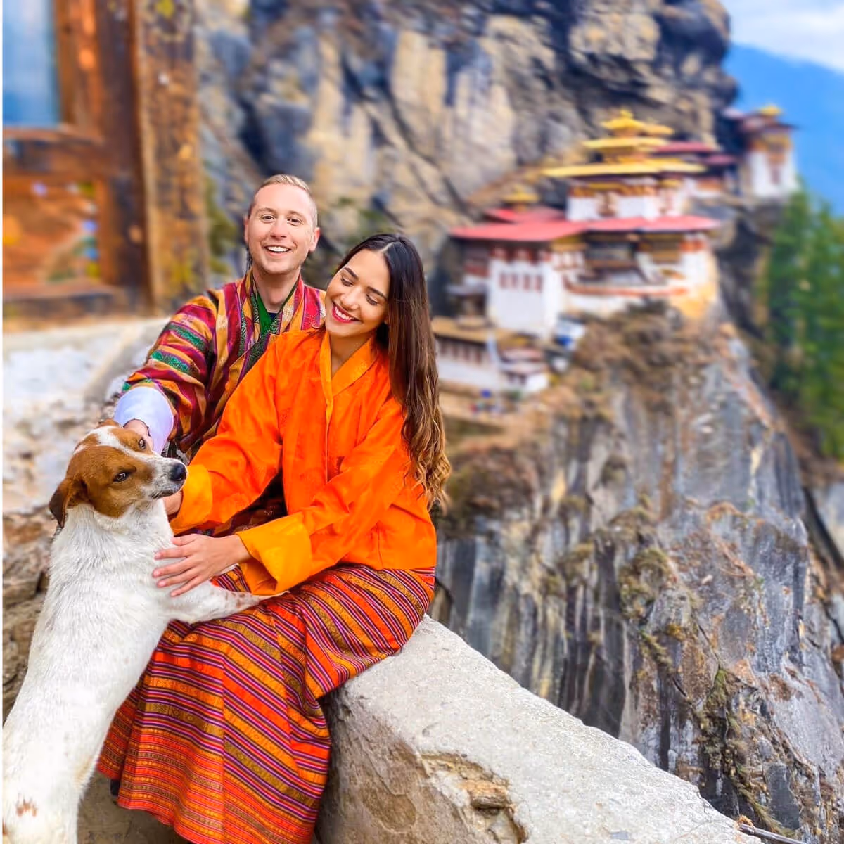 Happy couple in traditional Bhutanese attire with a dog, overlooking Tiger’s Nest Monastery in Paro.