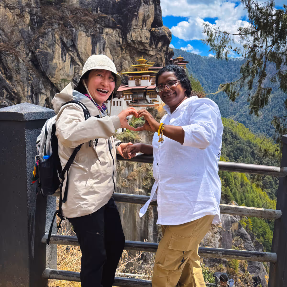 Two friends smiling and forming a heart shape with their hands in front of Bhutan’s iconic Tiger’s Nest Monastery, celebrating connection and adventure.
