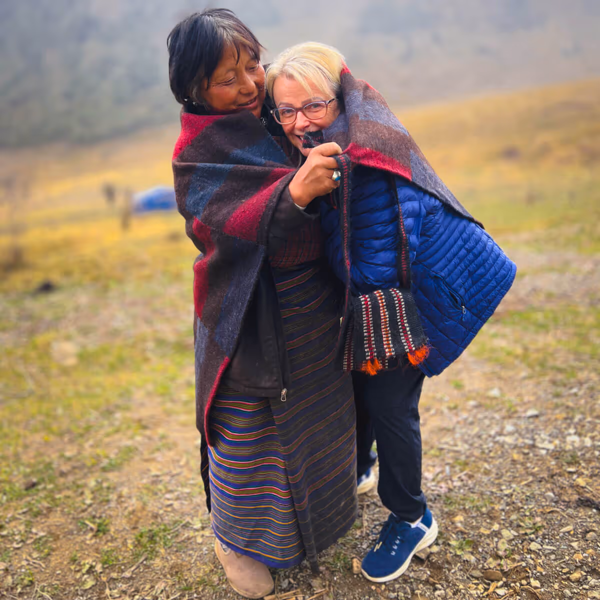 Two women warmly wrapped in a shared blanket, sharing a joyful moment in Bhutan’s highland meadows.