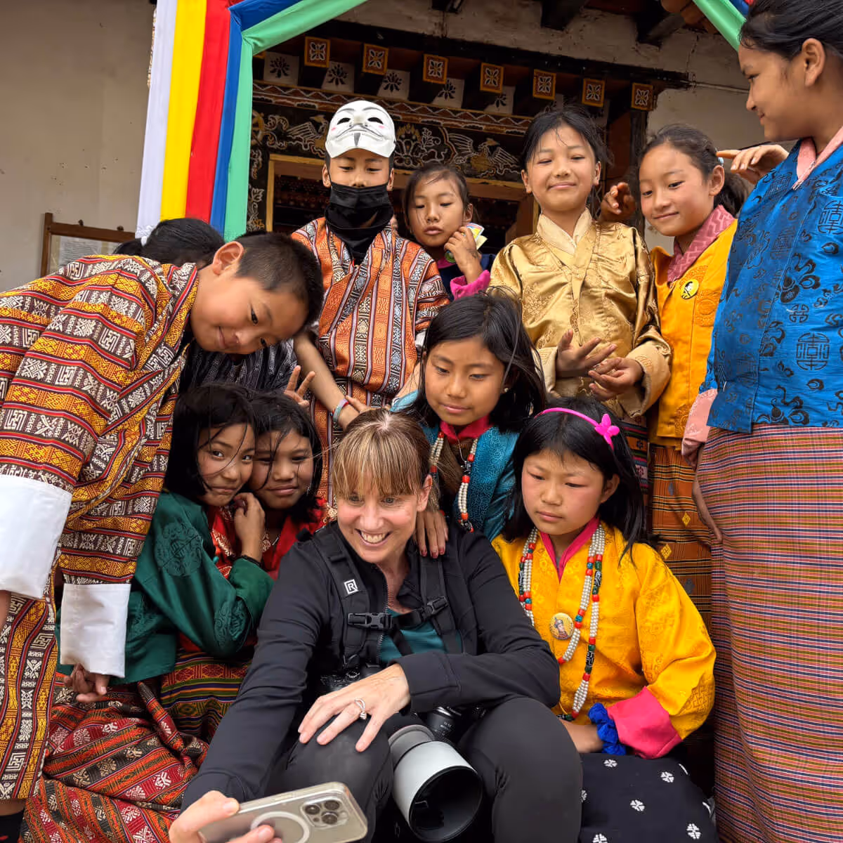Woman taking a selfie surrounded by Bhutanese children in vibrant traditional attire, capturing pure delight.