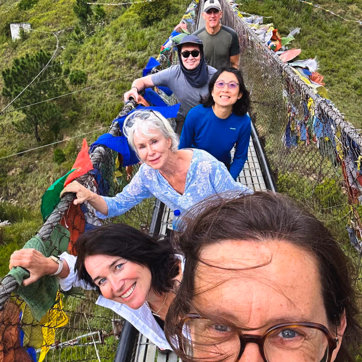 Group of smiling guests crossing a suspension bridge adorned with colorful prayer flags in Bhutan.