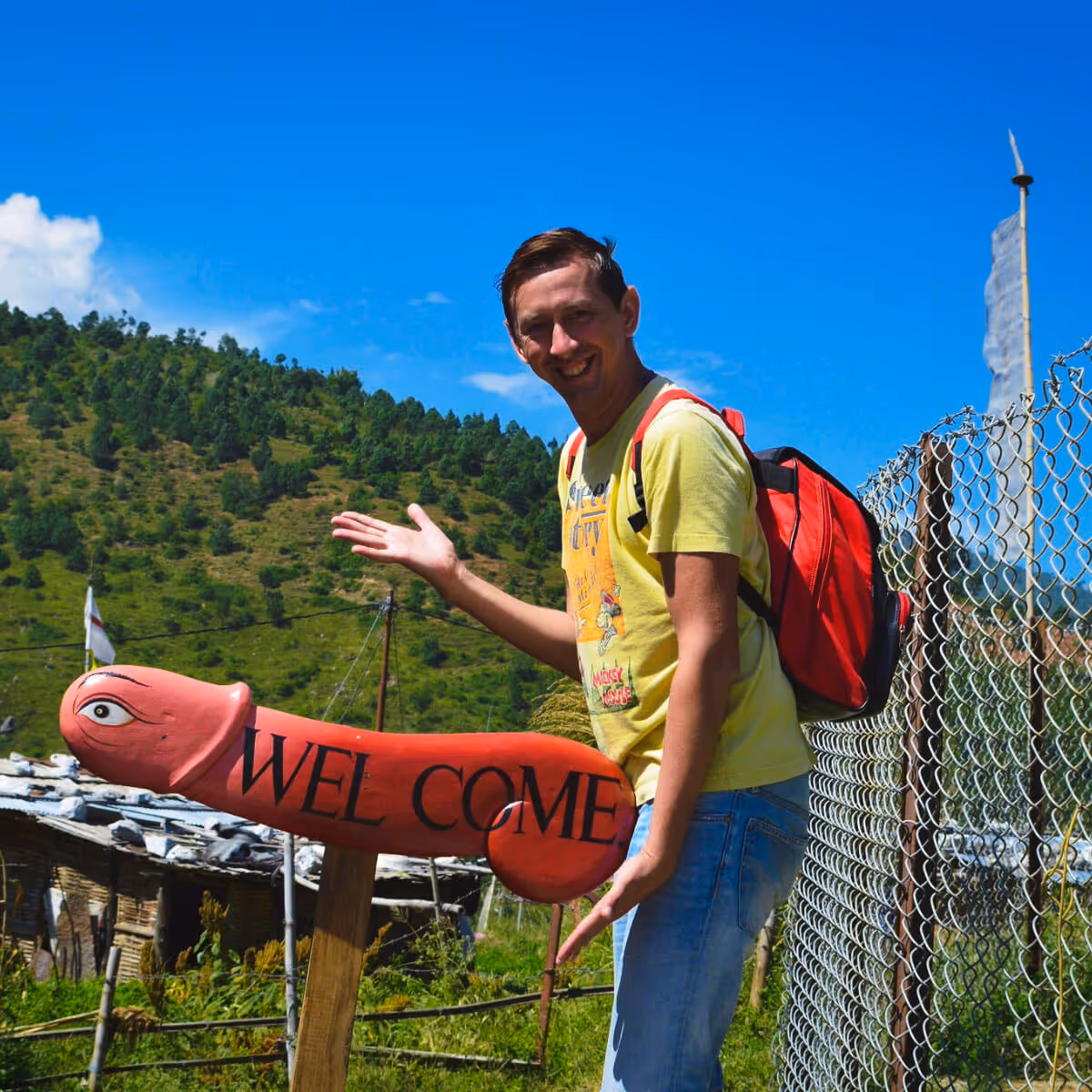 Guest posing playfully beside traditional phallus welcome sign in Bhutan, embracing local humor.