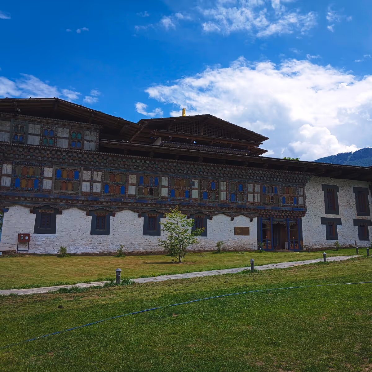 Front view of Wangdicholing Palace in Bumthang, Bhutan, featuring ornate painted windows, a green lawn, and a vivid blue sky.