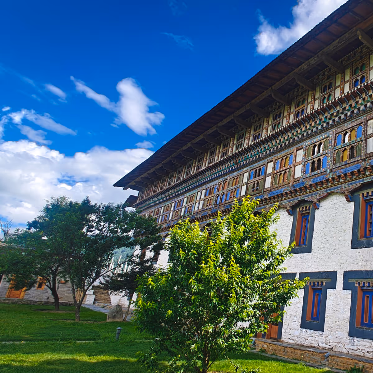 Ornate facade of Wangdicholing Palace in Bumthang, Bhutan, with traditional painted windows and lush green gardens beneath a bright blue sky.