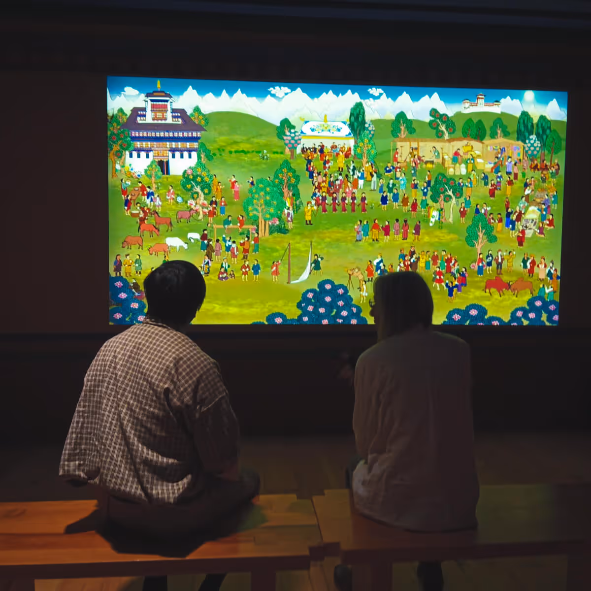 A guest seated with her guide inside Wangdicholing Palace in Bumthang, Bhutan, admiring a vibrant mural depicting local festival and village life.