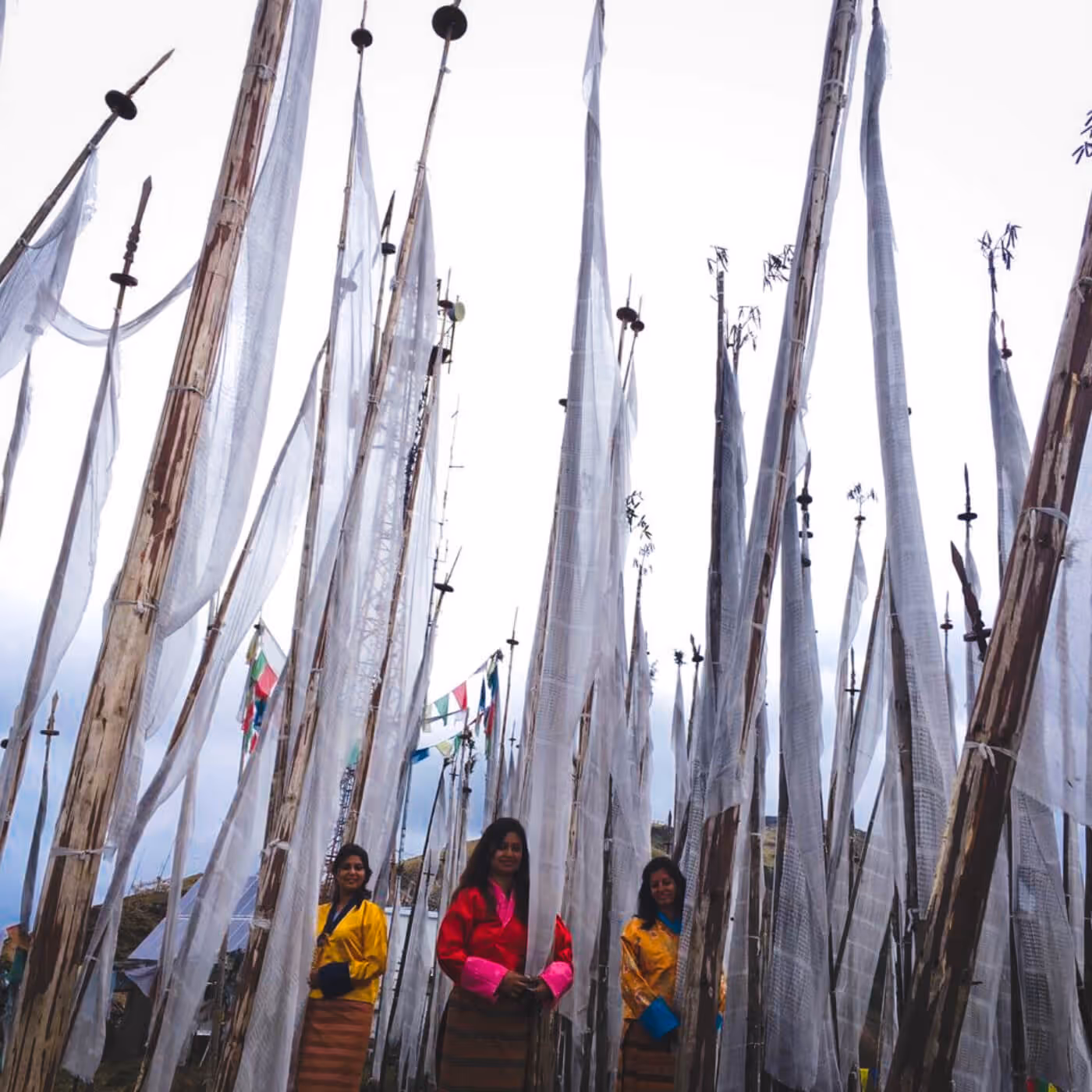 Three women in colorful traditional kira standing gracefully among tall white prayer flags at Chelela Pass.