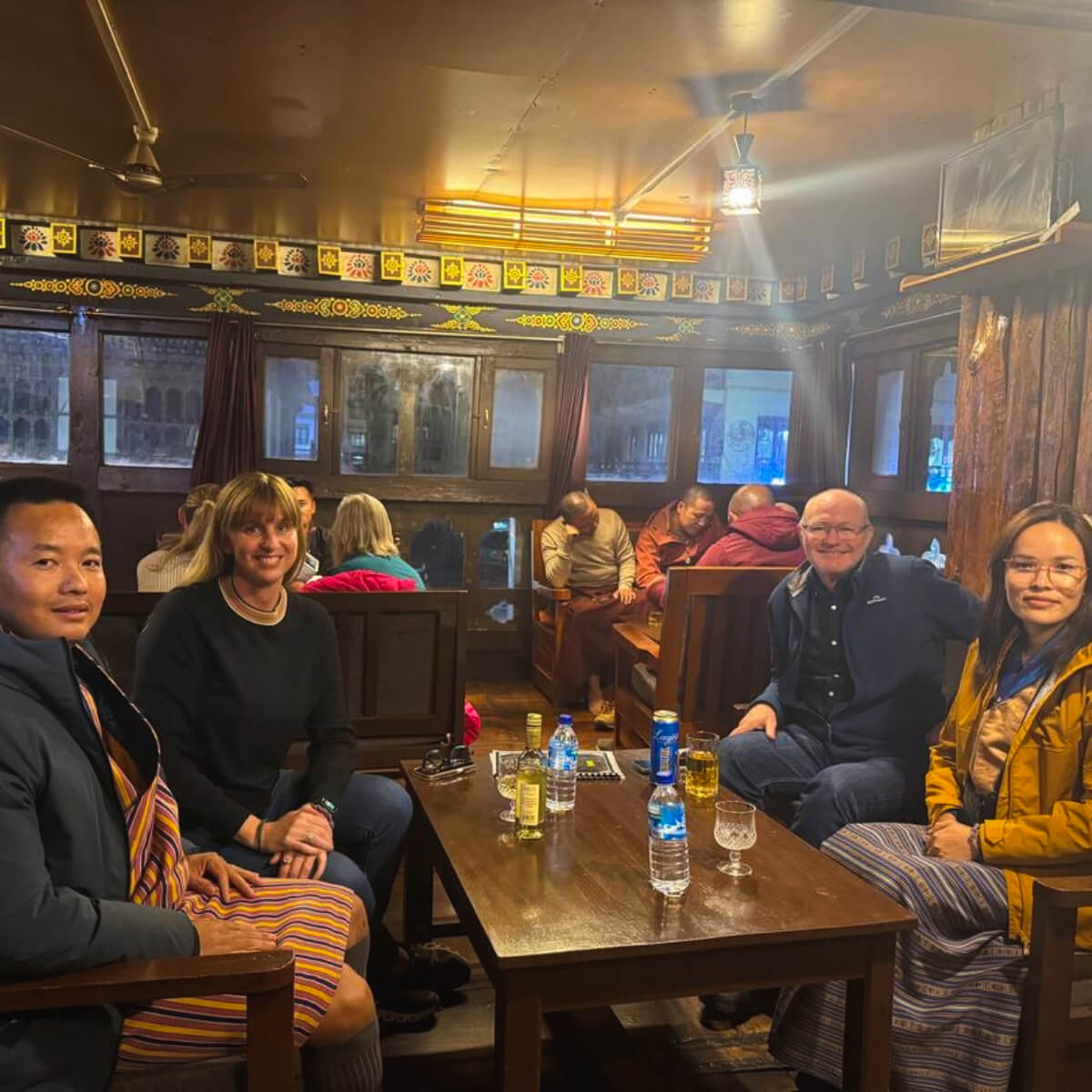Guests and guides enjoying drinks and warm conversation inside Kuzu Restaurant in Paro, Bhutan, surrounded by traditional wooden decor.