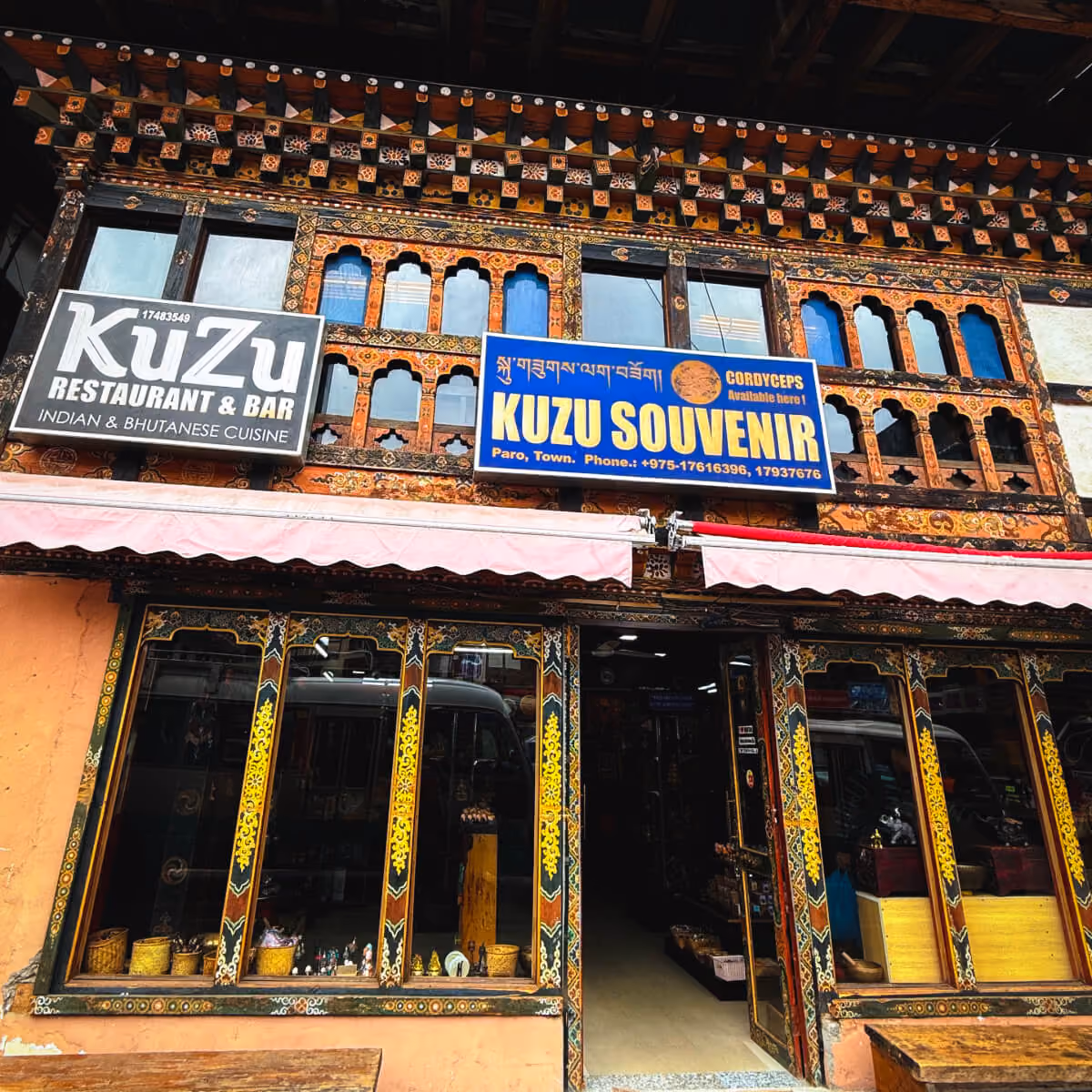 Exterior view of Kuzu Restaurant & Bar and Kuzu Souvenir shop in Paro, Bhutan, featuring intricate wooden window frames and vibrant storefront signage.