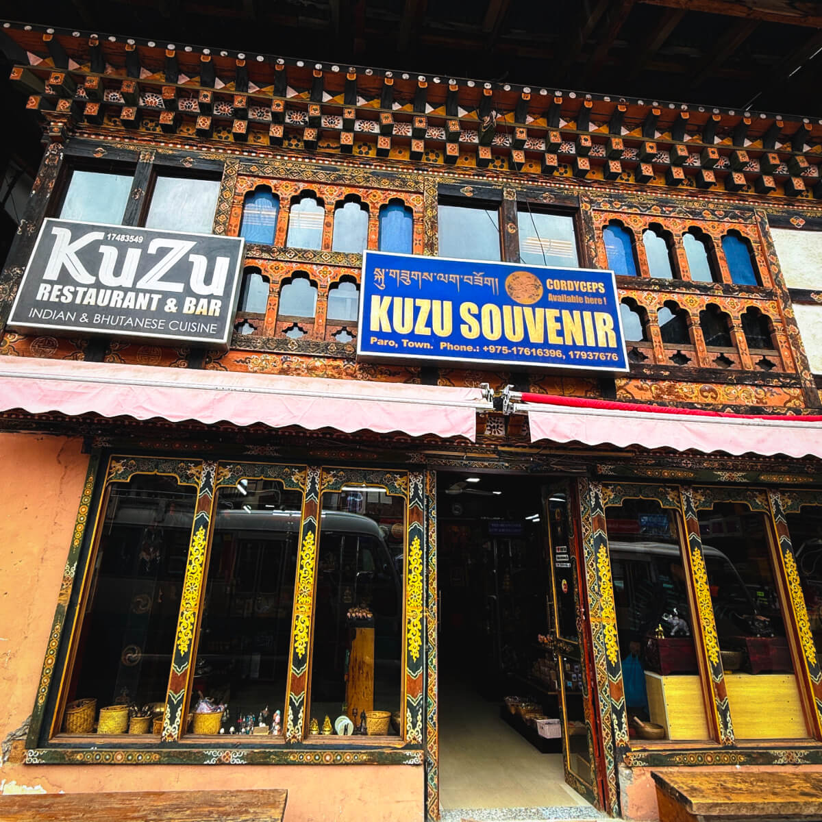Exterior view of Kuzu Restaurant & Bar and Kuzu Souvenir shop in Paro, Bhutan, featuring intricate wooden window frames and vibrant storefront signage.