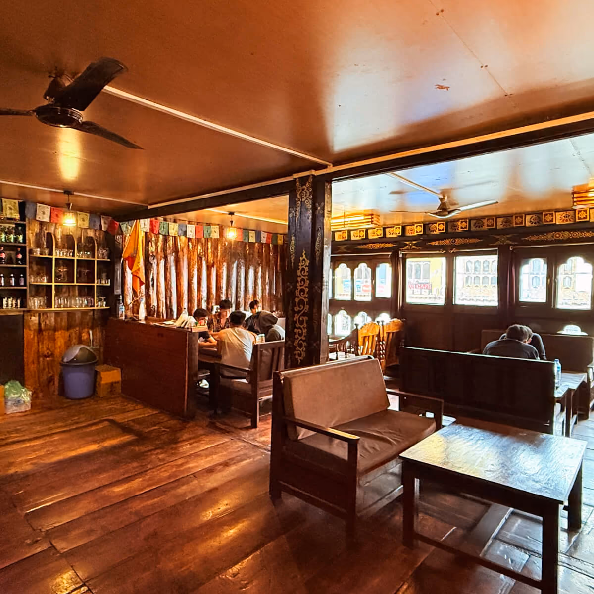 Cozy interior of Kuzu Restaurant in Paro, Bhutan, featuring wooden decor, prayer flags, and guests sharing meals in a warm, inviting atmosphere.