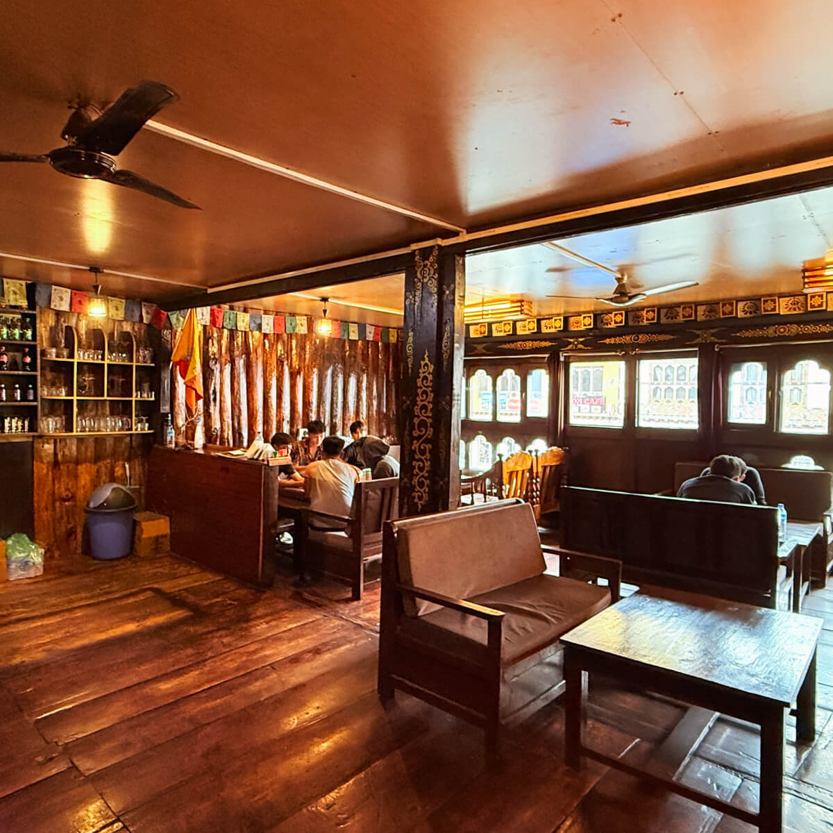 Cozy interior of Kuzu Restaurant in Paro, Bhutan, featuring wooden decor, prayer flags, and guests sharing meals in a warm, inviting atmosphere.
