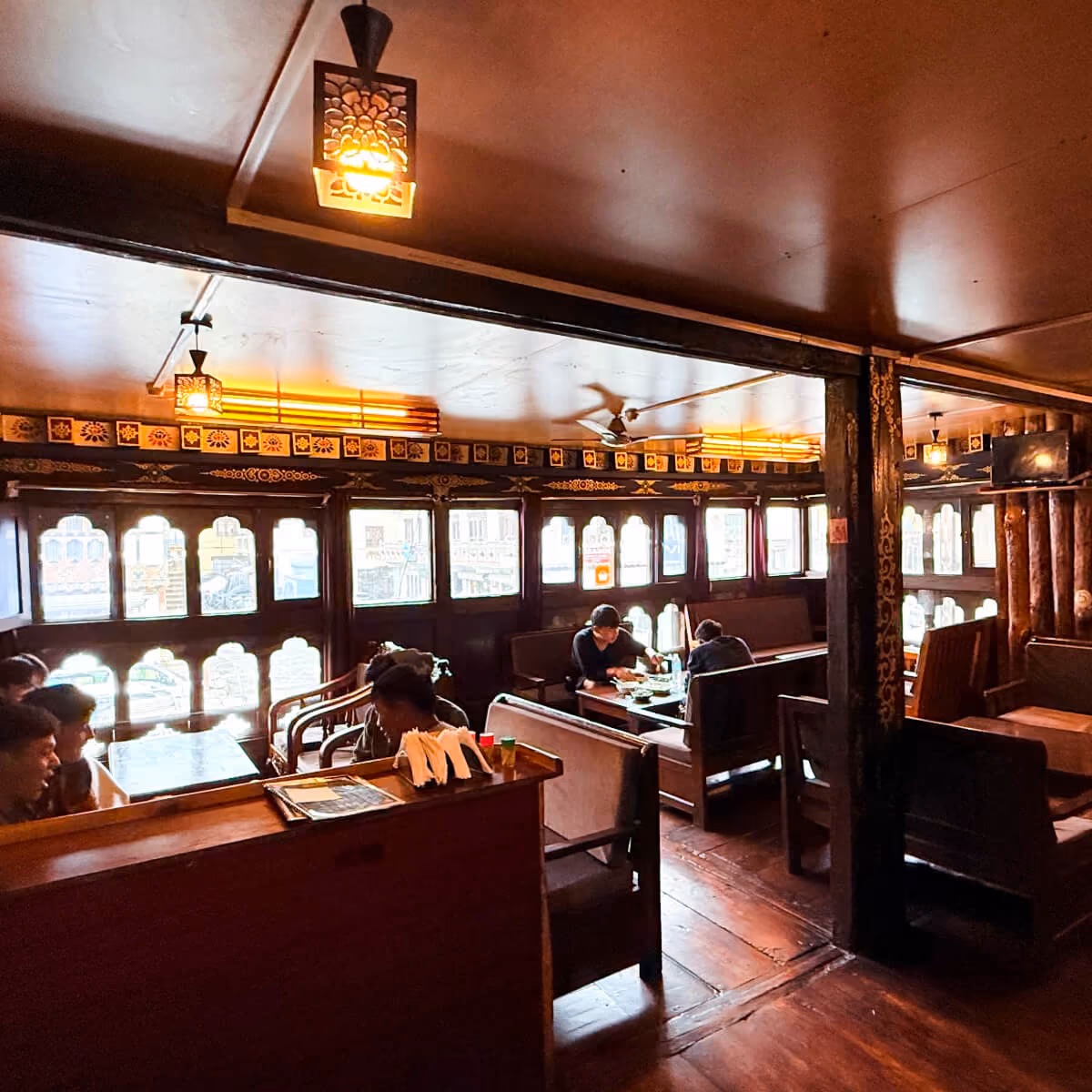 Warmly lit dining space at Kuzu Restaurant in Paro, Bhutan, where guests enjoy meals at wooden tables surrounded by traditional carved windows and decorative details.