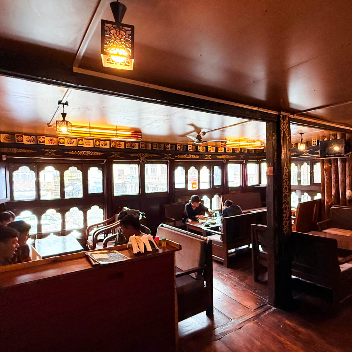 Warmly lit dining space at Kuzu Restaurant in Paro, Bhutan, where guests enjoy meals at wooden tables surrounded by traditional carved windows and decorative details.
