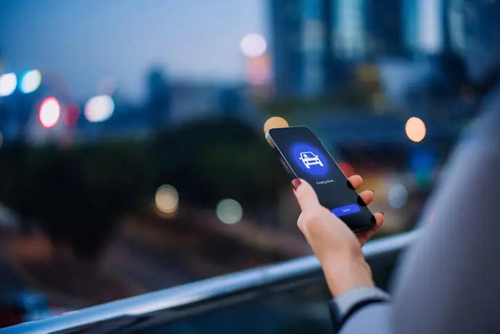 Une personne utilise une application mobile pour commander un véhicule, en soirée, depuis un balcon surplombant une ville moderne.